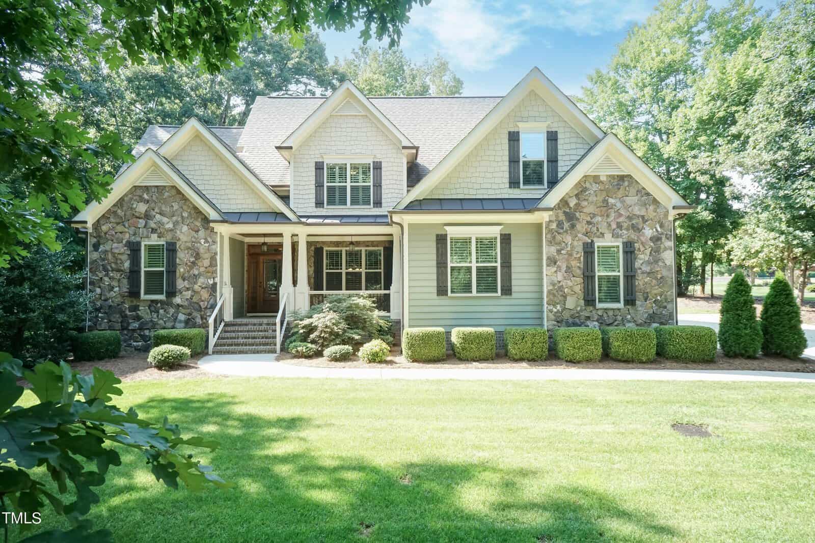 Nestled on 3047 E Bay Court, this charming two-story house in Creedmoor boasts stone and shingle siding, surrounded by lush greenery and shrubs. A well-maintained lawn stretches across the front, with a paved walkway leading to the entrance under the canopy of tall trees against a clear blue sky.
