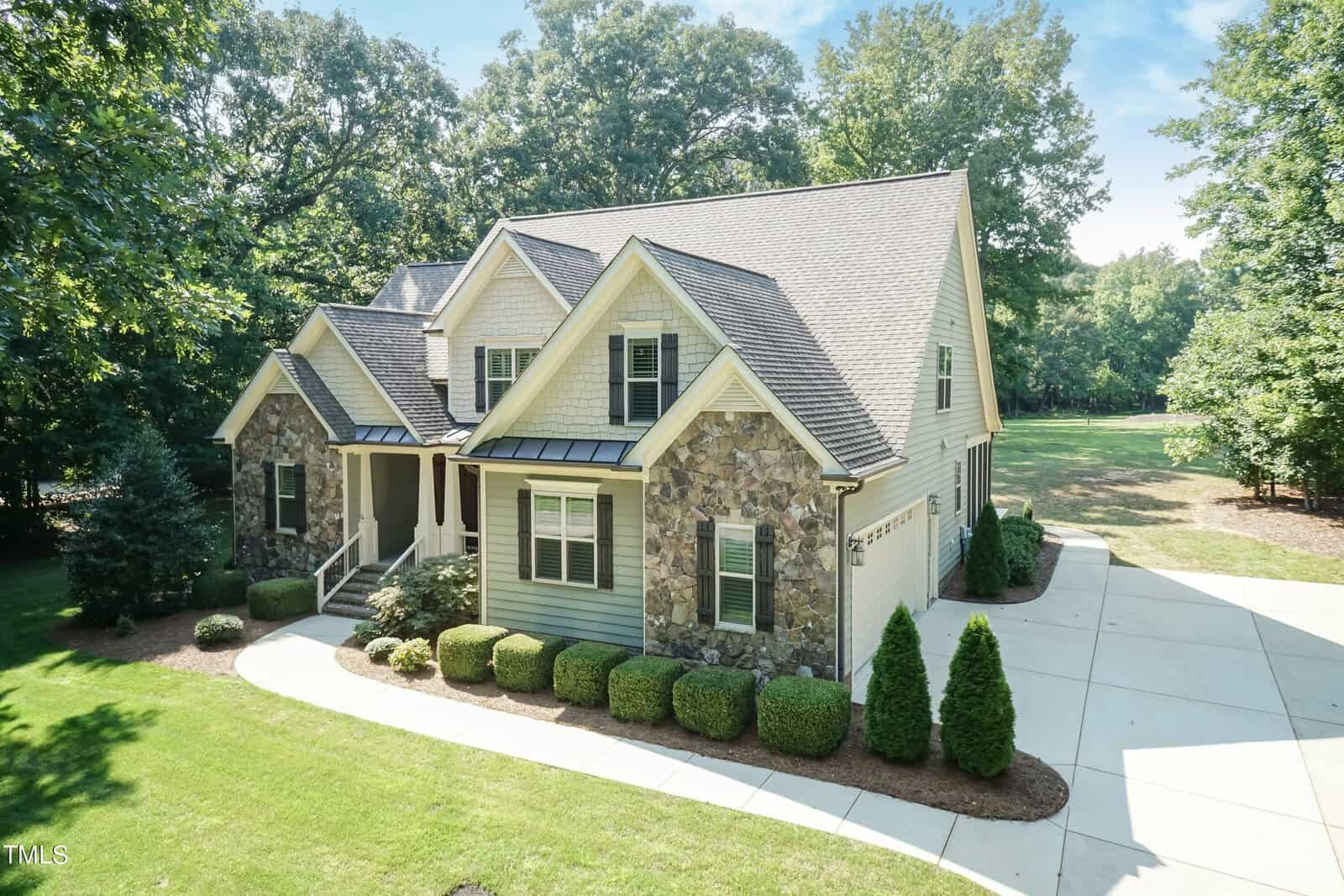 A stone and siding house with dormer windows and a porch, surrounded by lush greenery. The driveway is curved, and the property is bordered by tall trees under a blue sky.