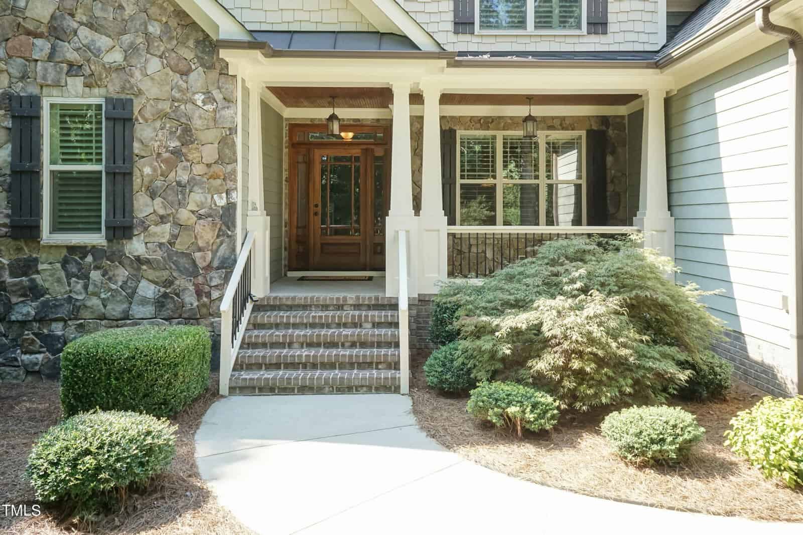 Front entrance of a home with a stone and wood facade. The covered porch has a wooden door with glass panels and white railings. Brick steps lead up to the porch, surrounded by manicured shrubs and bushes.