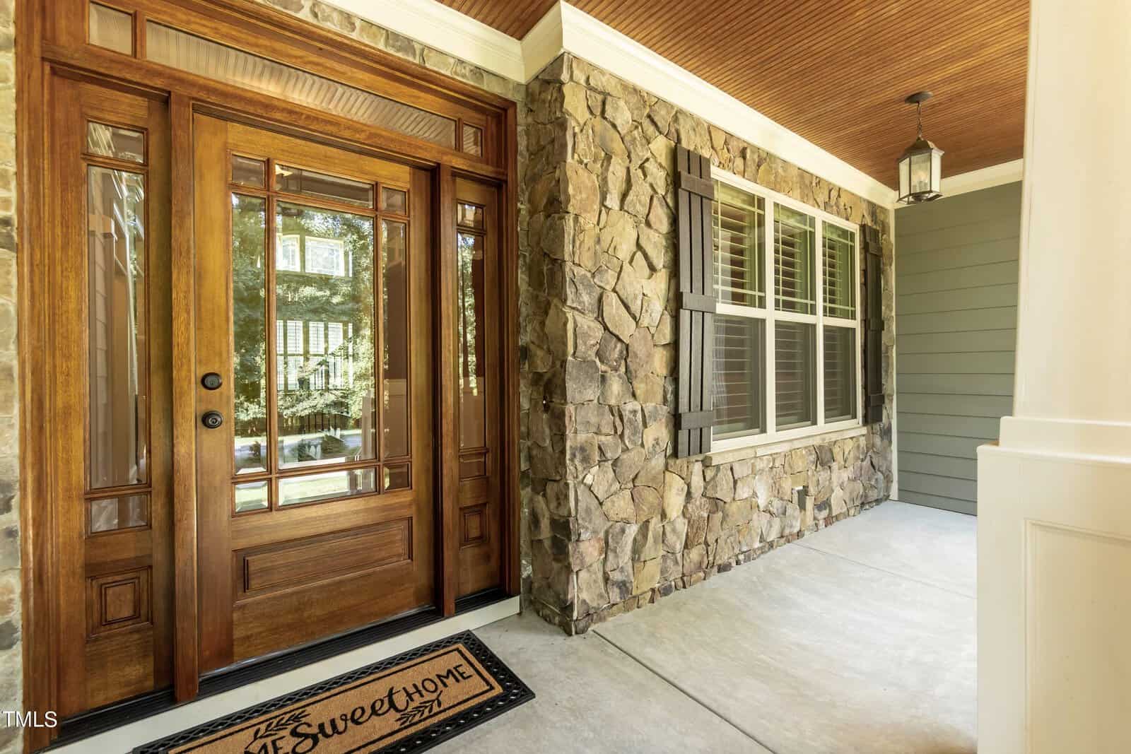 A welcoming front porch with a large wooden door featuring glass panels, stone wall siding, and a "Home Sweet Home" doormat. A window with shutters is nearby, and a light hangs from the wooden ceiling.