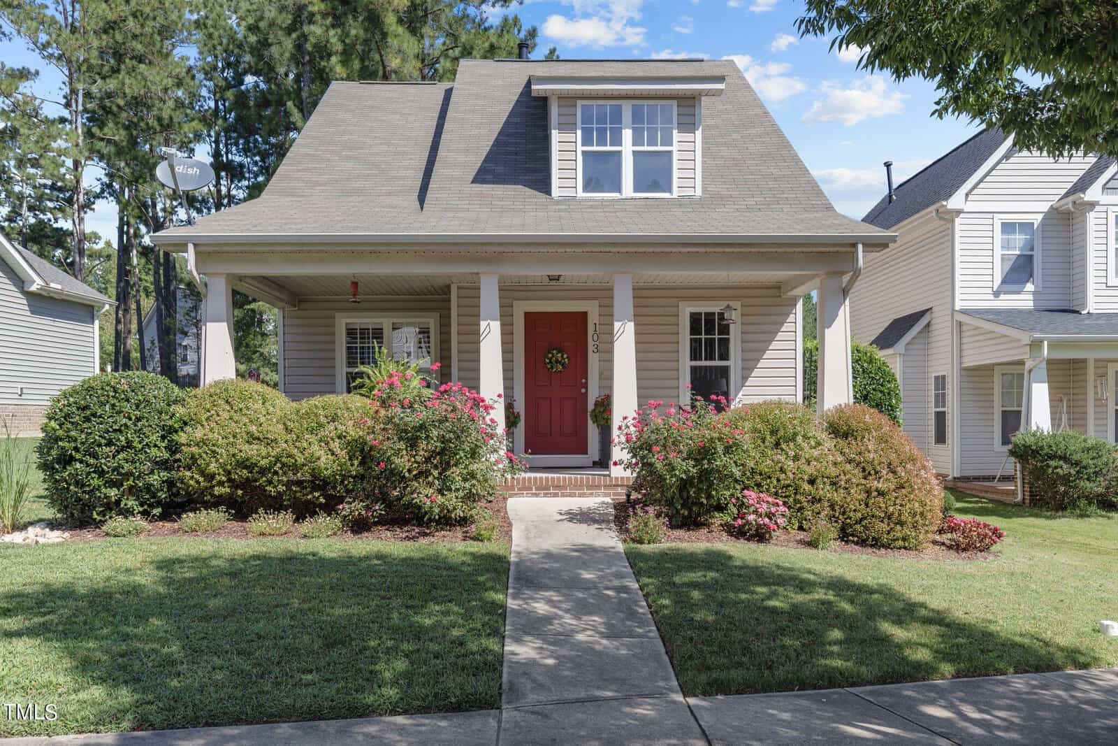 A quaint, single-story house at 103 Kipling Drive boasts a red front door, flanked by bushes and a manicured lawn. The porch features white columns and a wreath on the door. Nestled in Oxford, it is surrounded by greenery and neighboring homes under a clear blue sky.