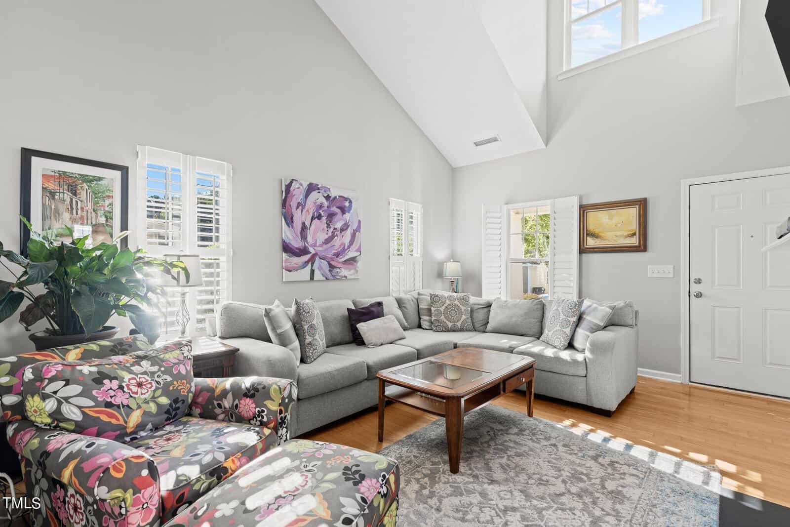 A bright living room with a large gray sectional sofa and a glass-topped coffee table. Two colorful floral armchairs face the sofa. Sunlight streams through plantation shutters, illuminating the hardwood floor and wall art, including a purple flower painting.