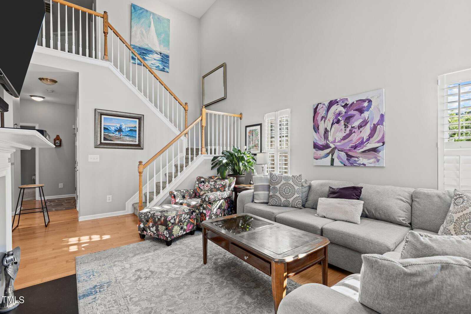 A bright living room featuring a gray sectional sofa with patterned pillows, a floral armchair, and a wooden coffee table. Artworks adorn the light gray walls, and a staircase with wooden railings is visible in the background. Natural light fills the room.