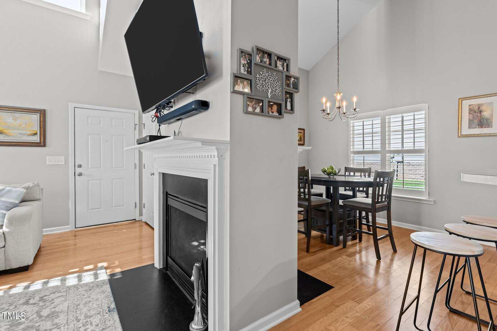 A living room area with a wall-mounted TV above a fireplace, and a dining area with a wooden table and chairs under a chandelier. The room has hardwood floors, light gray walls, and several windows allowing natural light.