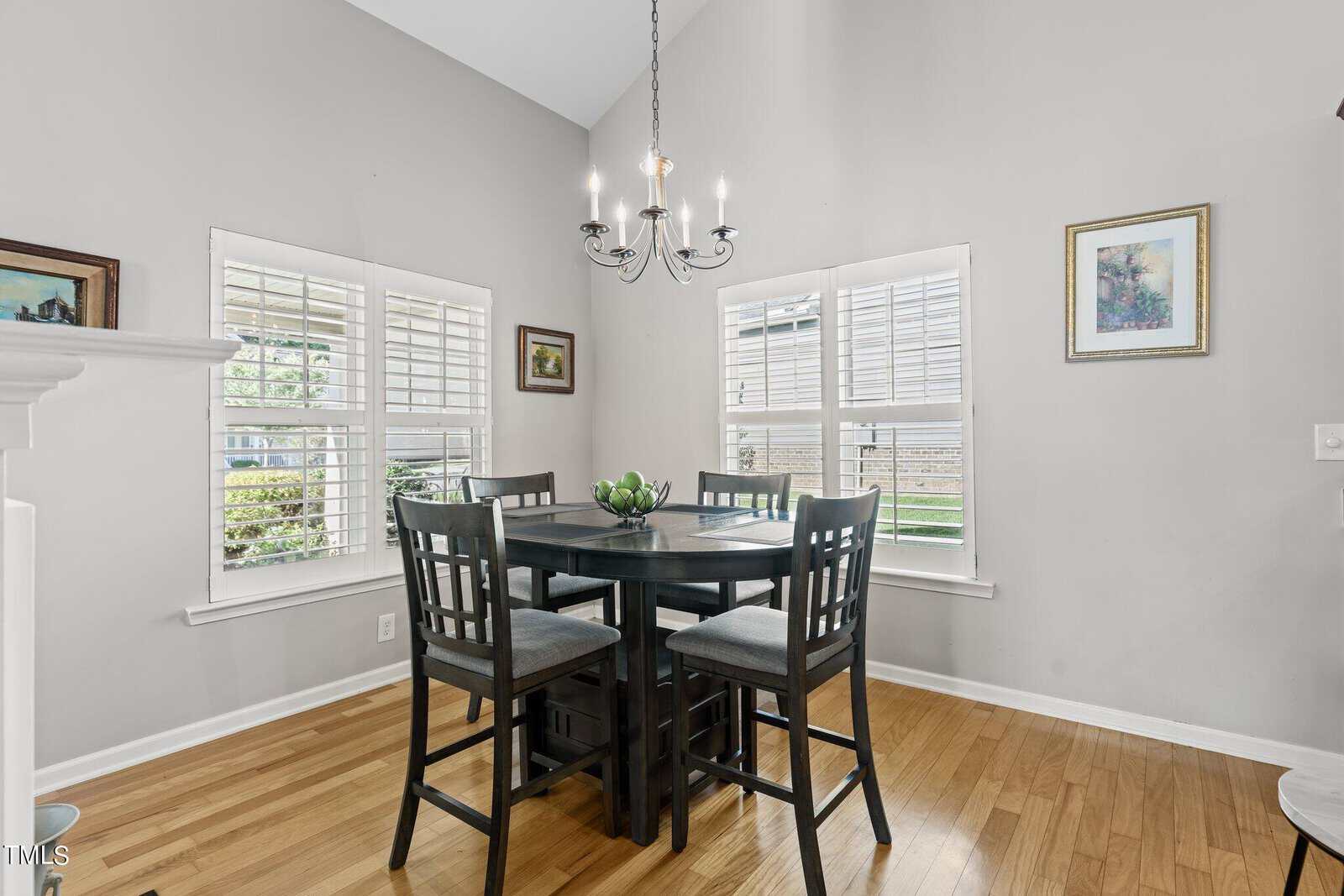 A bright dining area features a dark wooden table with four matching chairs. Large windows with white shutters let in natural light. A modern chandelier hangs above the table, and framed art decorates the light gray walls.