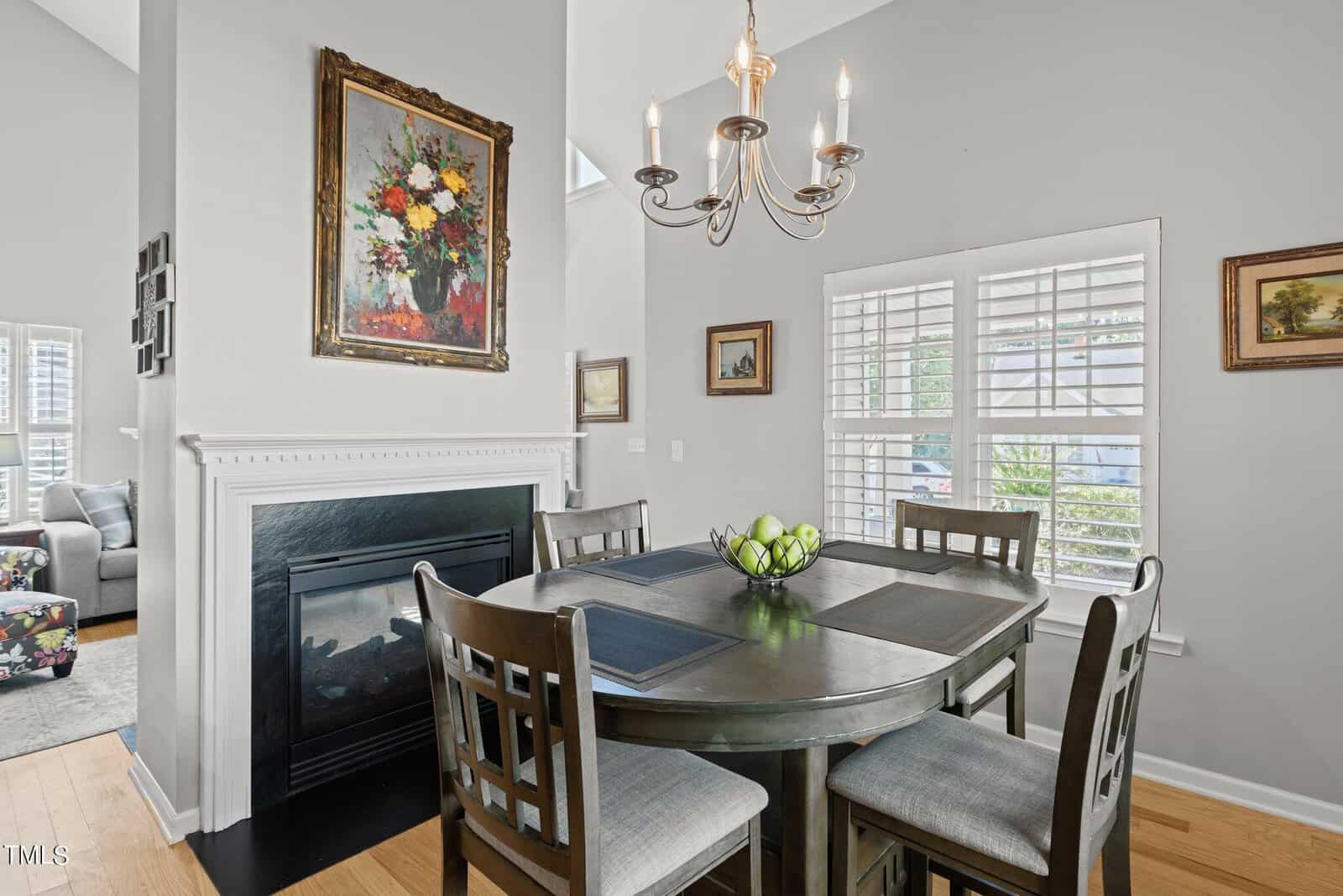 A dining area with a dark wood table set for four, featuring a bowl of green apples. A fireplace with a floral painting above it is in the background, under a chandelier. The space is brightened by natural light from windows with white shutters.