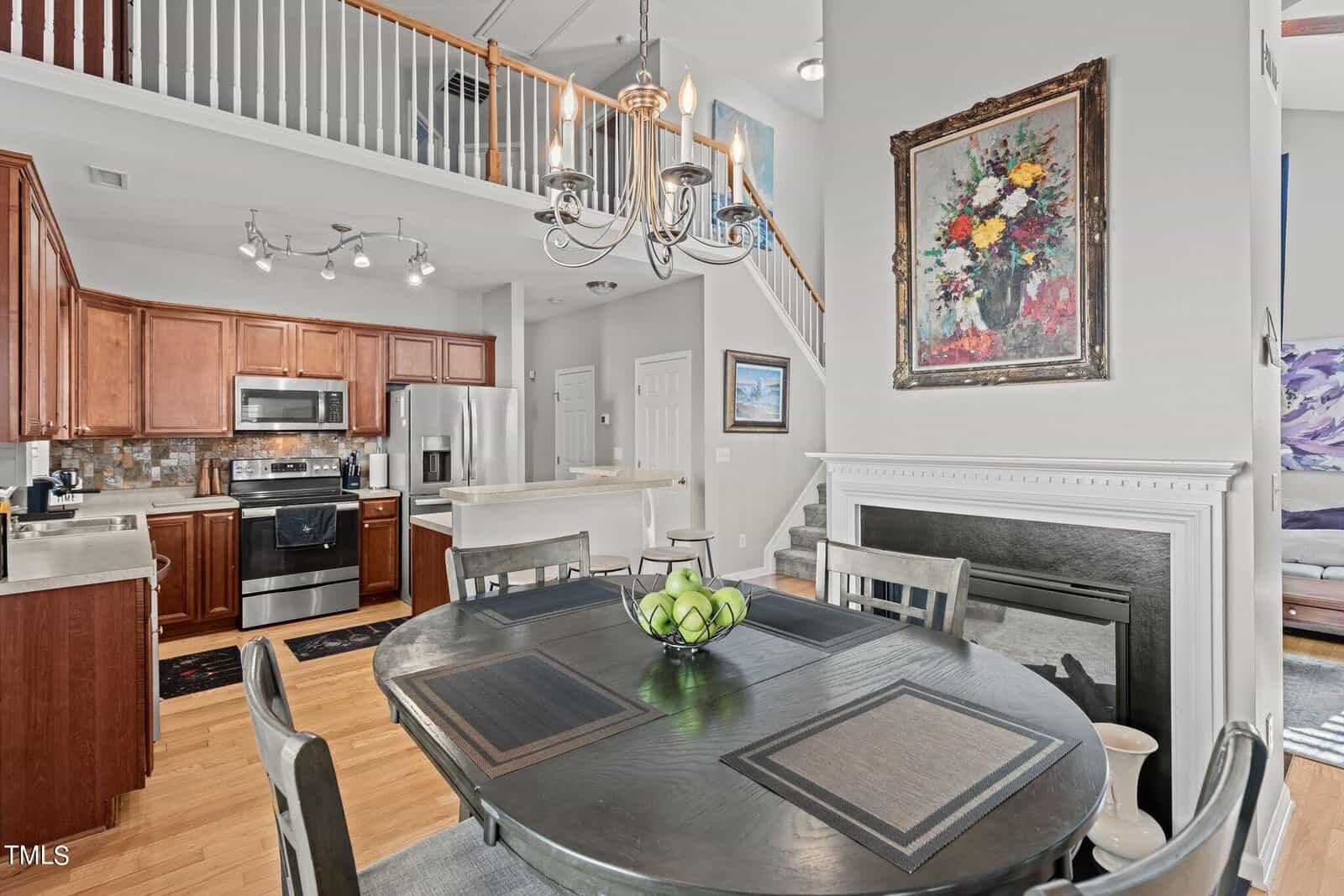 A modern kitchen and dining area with wooden cabinetry, stainless steel appliances, and a round dining table with a bowl of green apples. There's a chandelier, a fireplace, a floral painting, and a staircase leading to a loft above.