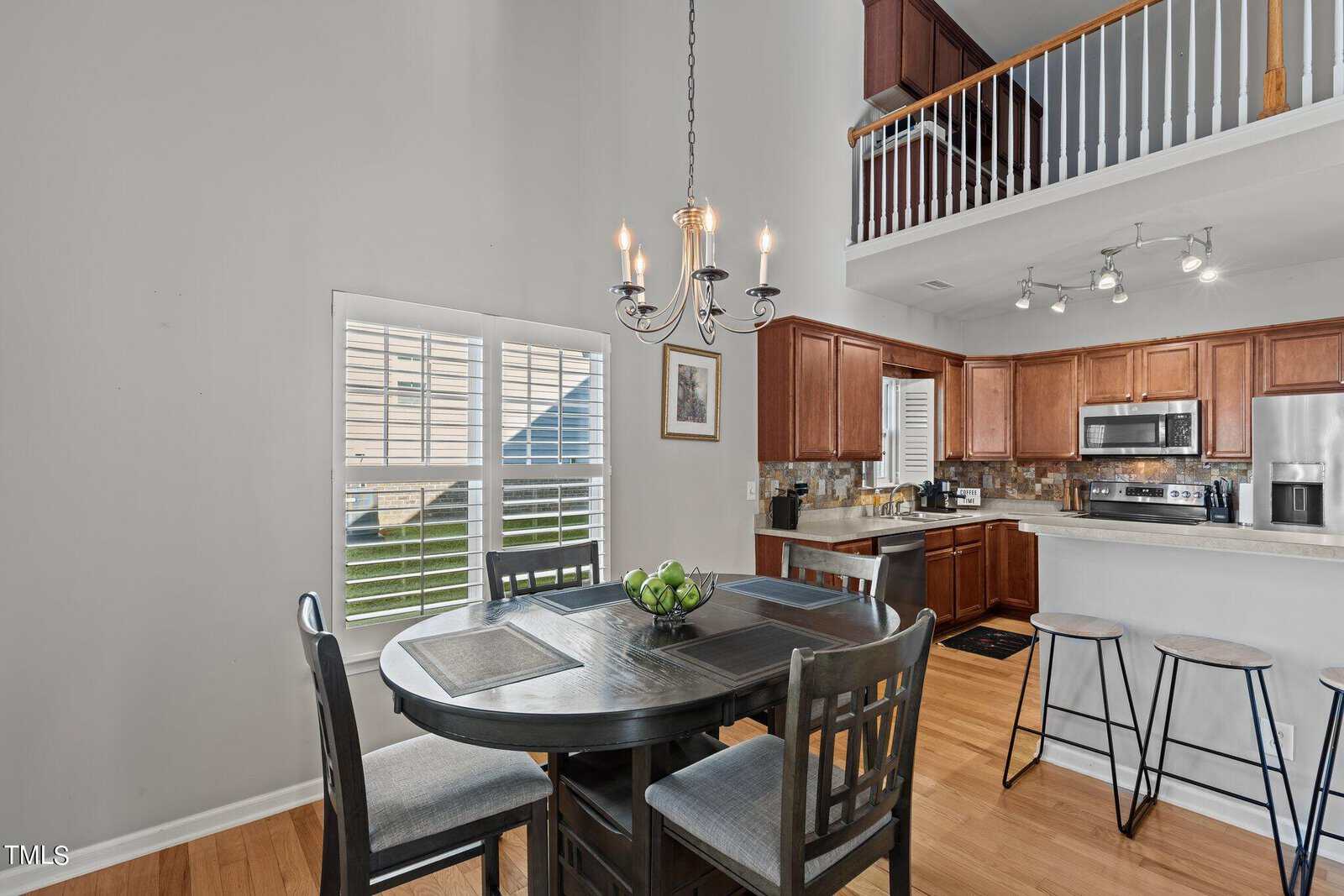 A modern kitchen and dining area with high ceilings. There's a dark wooden dining table with six chairs and a bowl of green apples. The kitchen features brown cabinets, stainless steel appliances, and a small bar area with stools. A chandelier hangs overhead.