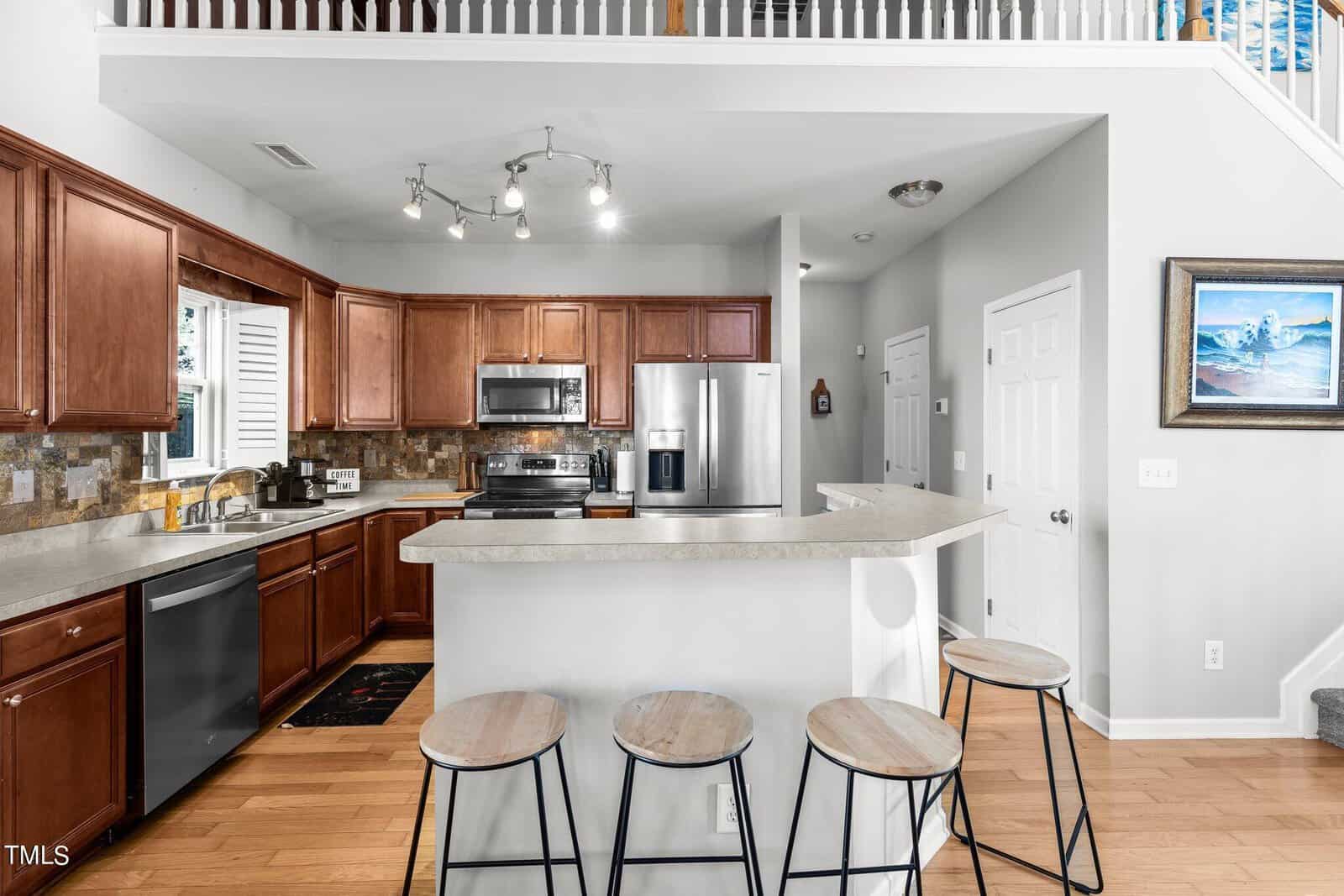 Modern kitchen with wooden cabinets and light countertops. A central island features bar stools. Stainless steel appliances, including a refrigerator and oven, are present. Overhead, track lighting provides illumination, and there is a staircase to the right.