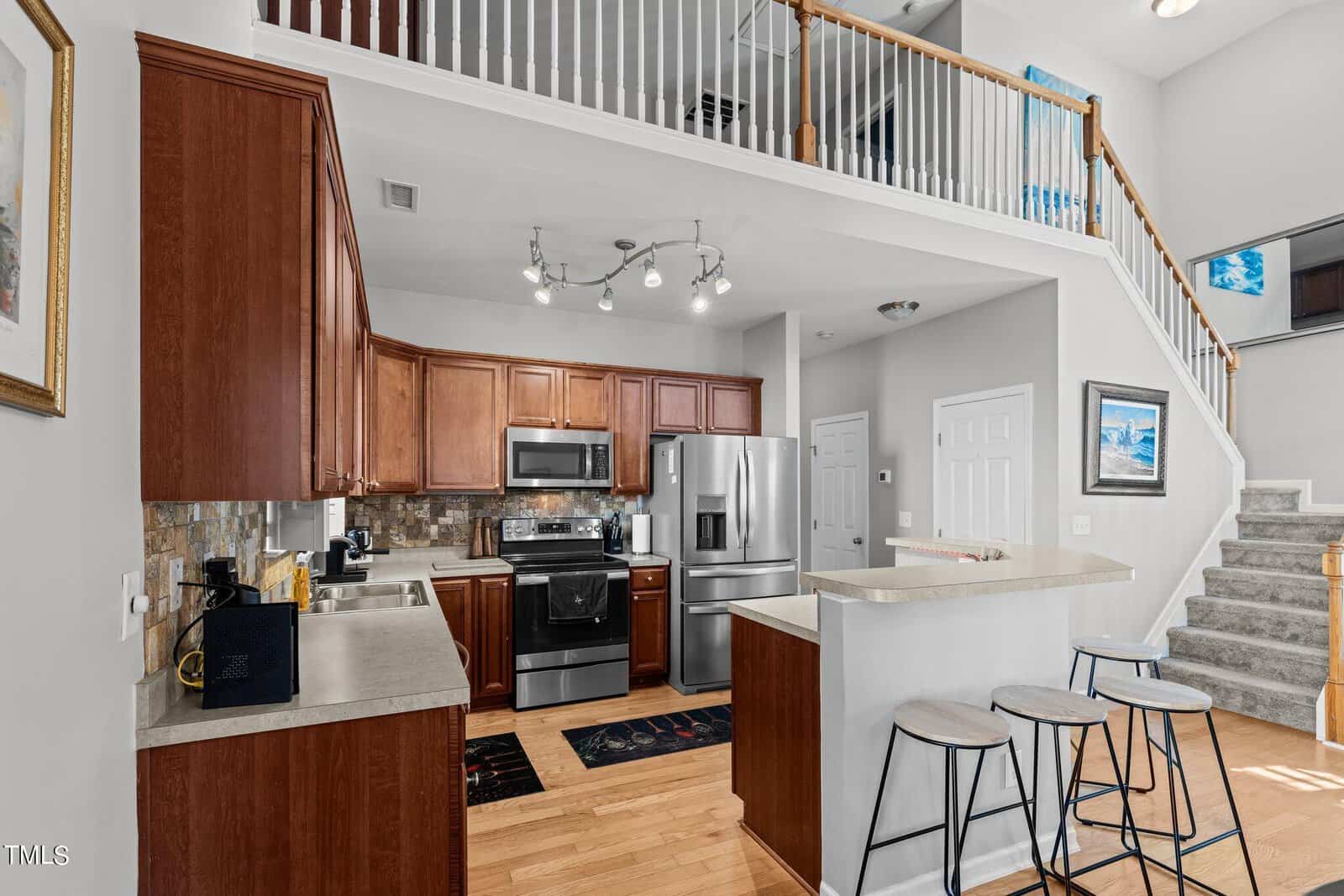 Modern kitchen with wooden cabinets, stainless steel appliances, and a breakfast bar with three stools. Overhead lighting illuminates the space. Stone backsplash and wooden floors add warmth. A staircase with white railing leads to an upper floor.