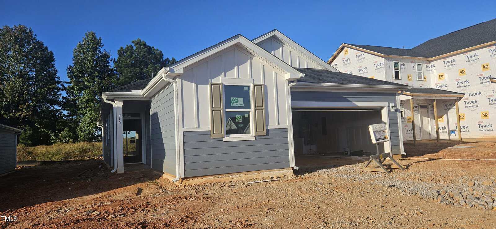 A single-story house under construction with gray siding, white trim, and a two-car garage. Windows have stickers, and the ground in front is unfinished dirt and gravel. Another house is partially visible next door.