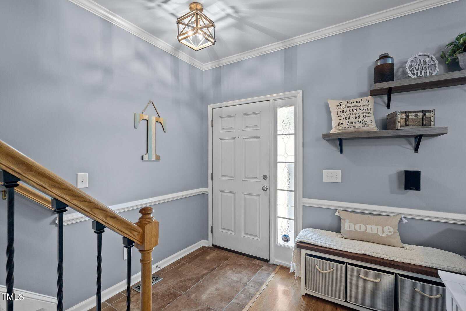 A home entryway with light blue walls and wooden flooring. There's a staircase with black railing on the left, a bench with storage bins and "home" pillows on the right, and a wall shelf with decor. A geometric light fixture hangs from the ceiling.