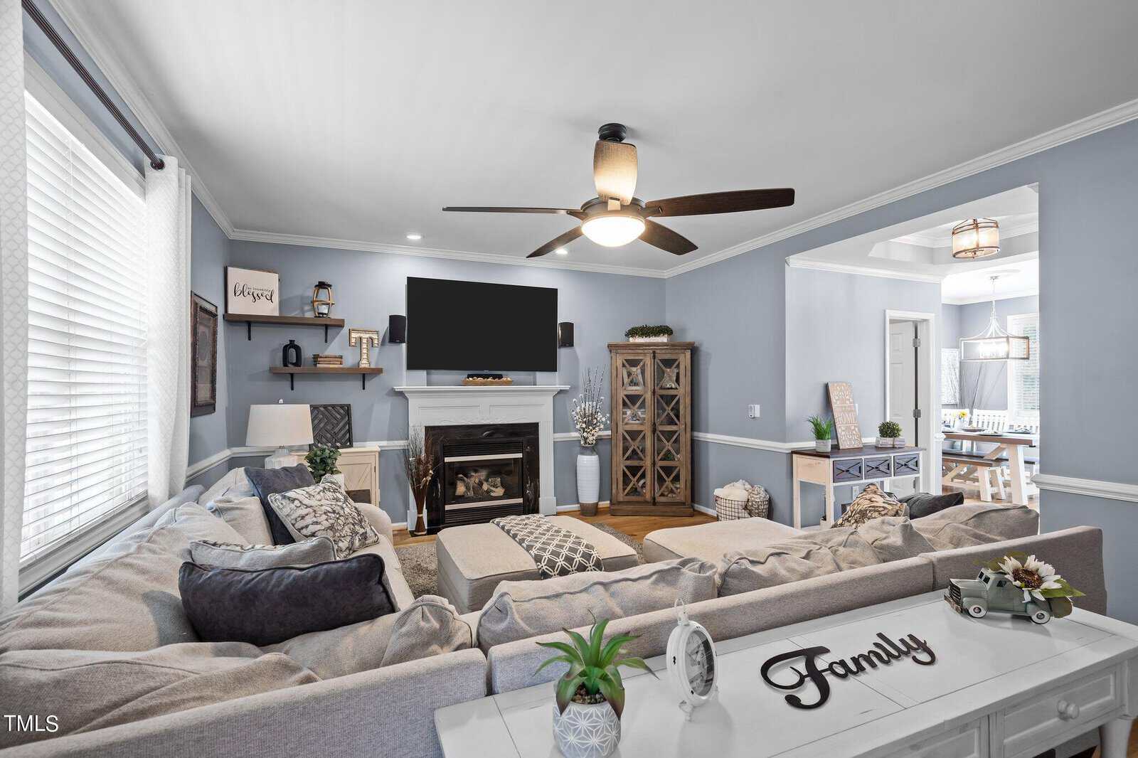 A cozy living room with a large gray sectional sofa facing a TV above a fireplace. The walls are light blue, and there's a ceiling fan. Decorations include a "Family" sign, plants, and shelves. The dining area is visible in the background.