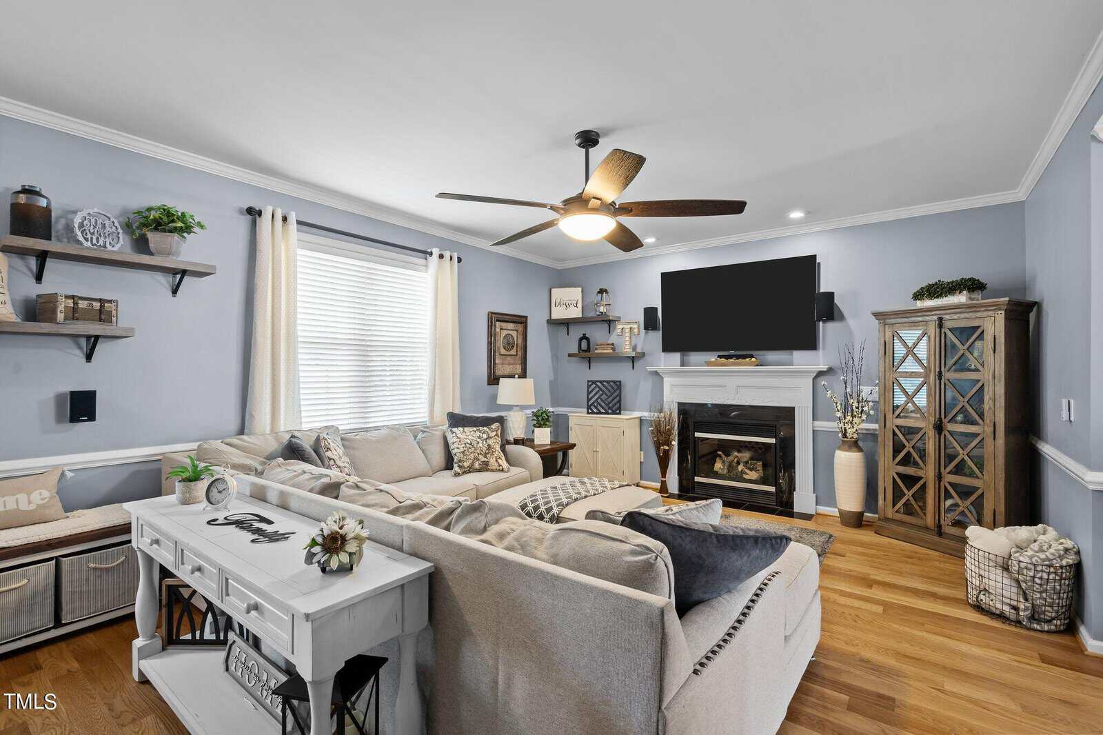 A cozy living room with light blue walls, featuring a large gray sectional sofa, a white fireplace with a TV above it, and a ceiling fan. There are decorative shelves, a cabinet, and a soft rug on the wooden floor. Natural light fills the room.