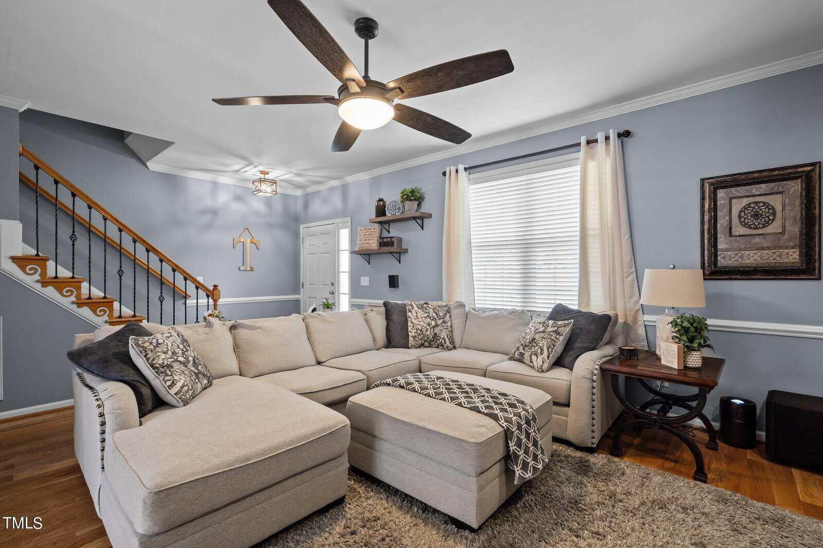 A cozy living room with a large beige sectional sofa and patterned cushions. A wooden coffee table holds a lamp and decor. The room features a ceiling fan, light blue walls, a staircase, and a window with blinds. A woven rug covers the hardwood floor.
