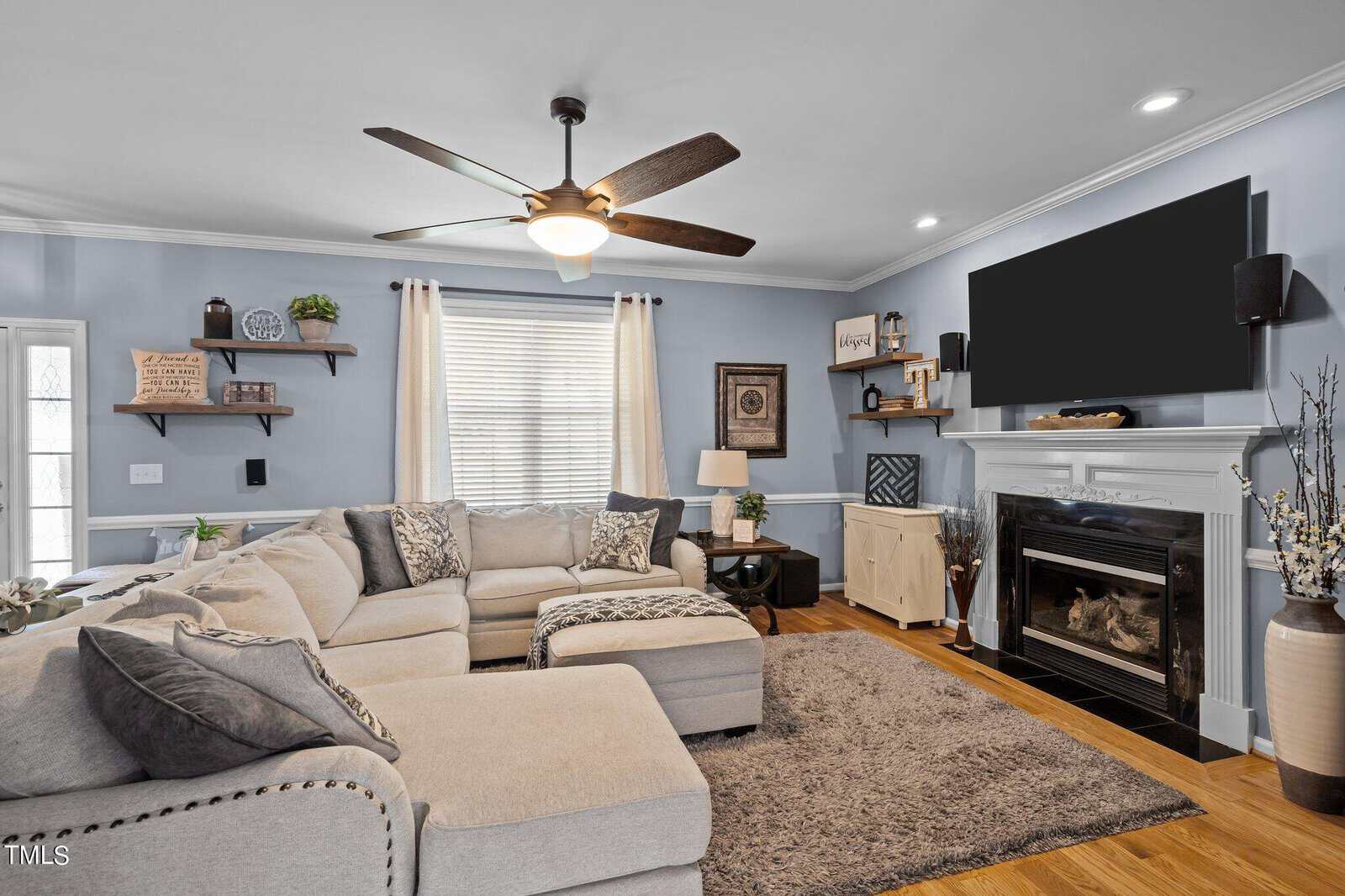 A cozy living room with a large beige sectional sofa and a shaggy rug. The walls are painted light blue, featuring a flat-screen TV above a white fireplace. Decorative shelves and framed art are on the walls, with a ceiling fan overhead.