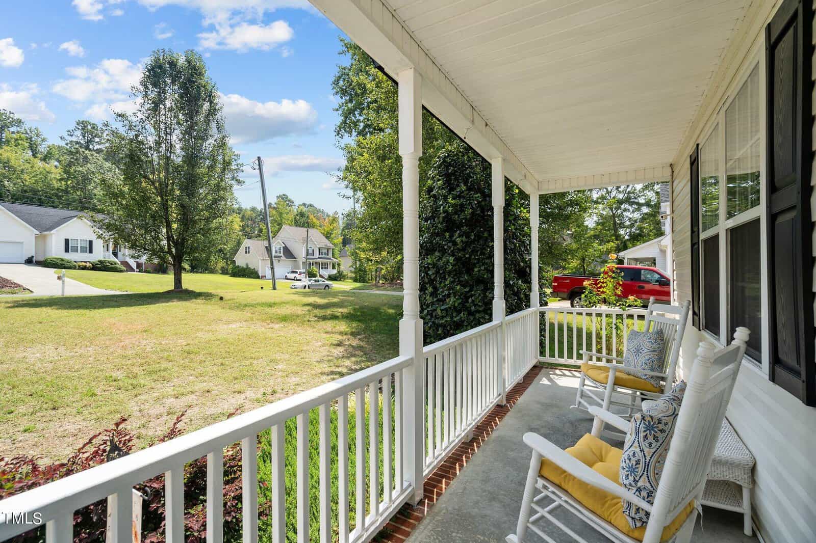A covered porch with two white rocking chairs and yellow cushions overlooks a suburban neighborhood. The lawn is green, and houses are visible in the background under a blue sky with white clouds.