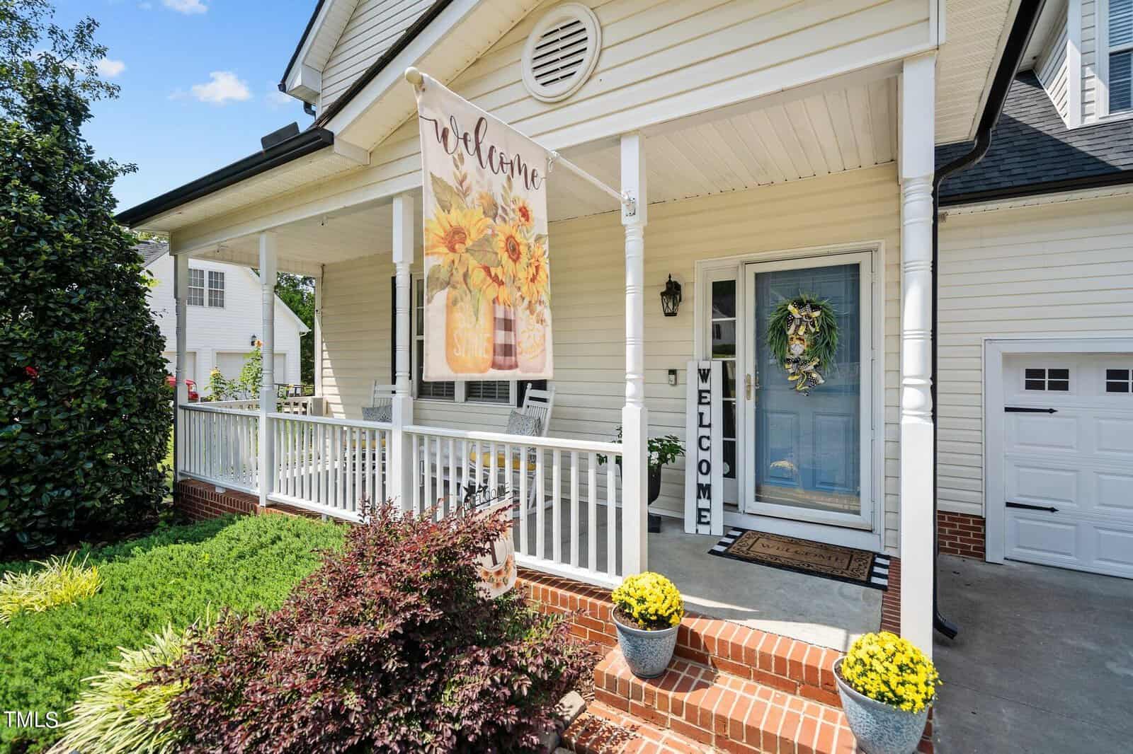 A charming house with a welcoming front porch displays a sign with sunflowers that reads "Welcome." The porch features a wreath on the door, a welcome mat, and potted yellow flowers on either side of the steps. A bright, sunny day completes the scene.