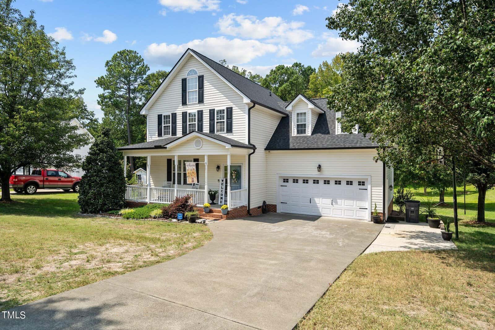 A two-story white house with black shutters and a front porch is surrounded by trees and a lawn. A concrete driveway leads to a two-car garage. The sky is clear with a few clouds. A red truck is parked on the lawn.