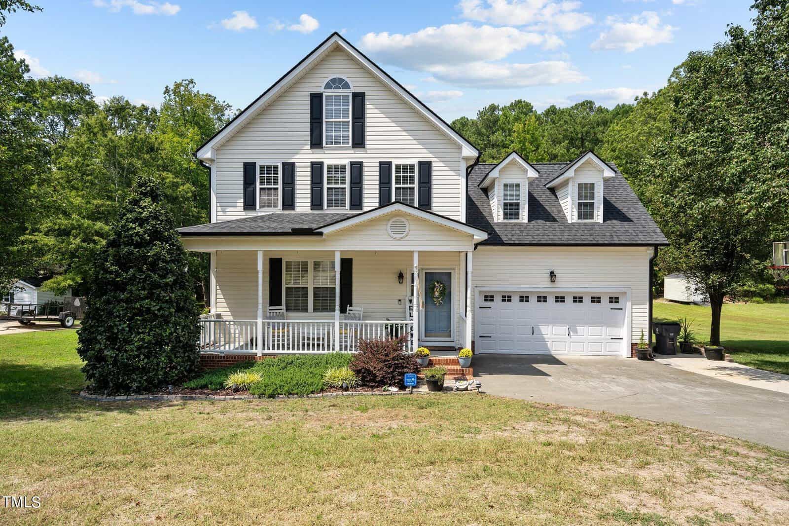 A two-story white house at 110 Oak Road in Butner boasts a black roof and a porch with white railings, complemented by a neatly maintained front yard. The closed garage door leads to a clean, spacious driveway, while tall trees provide a scenic backdrop.