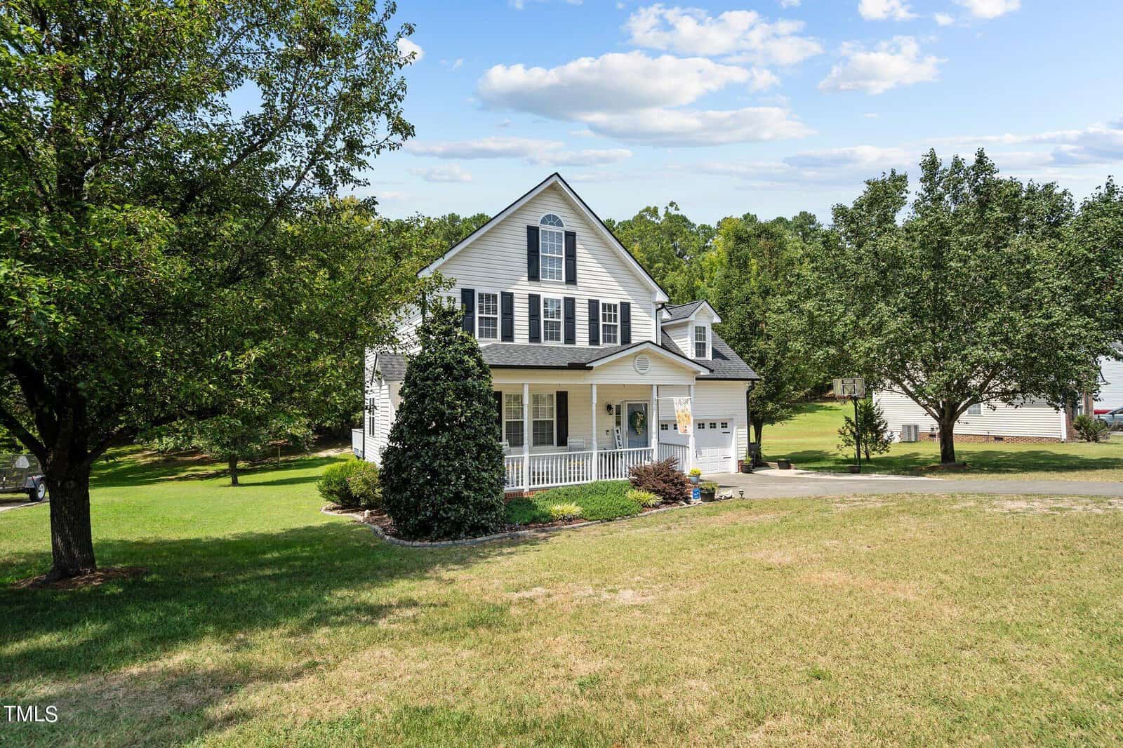 A two-story white house with black shutters and a front porch, surrounded by a well-maintained lawn and several trees. A driveway leads to a double garage. The sky is partly cloudy.
