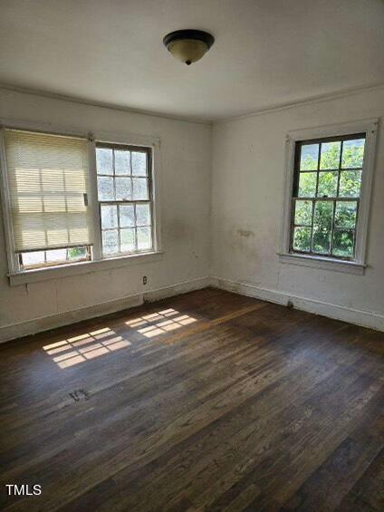 An empty room with two large windows allowing natural light to fill the space. The walls are partially discolored, and the wooden floor has visible marks and wear. A ceiling light fixture is centrally mounted. The view outside shows greenery.