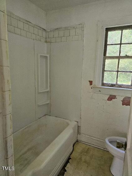An old bathroom with a discolored bathtub, dirty walls, and peeling paint. There is a window with cracked panes on the right and a white toilet in the back right corner. The tiles around the bathtub are also in poor condition.