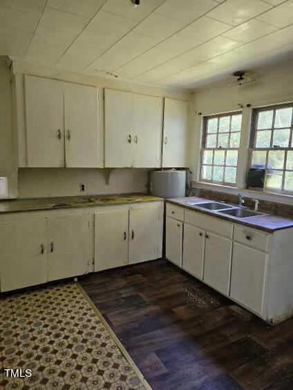 A small kitchen with white cabinets, a double sink beneath two windows, and a countertop with a few items. The floor has a patterned vinyl section and wood-like flooring. The light fixture on the ceiling looks dated, and the overall space appears somewhat worn.