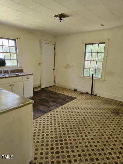 A dilapidated, empty room with worn, patterned flooring, partially missing in one area, revealing dark wooden planks. The walls are discolored with peeling paint. There are two windows and a door, and a small section of countertop with a sink and cabinets.