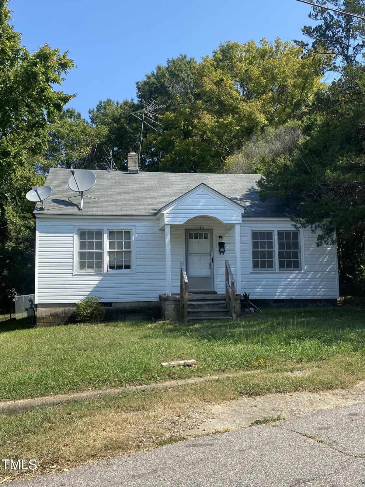 A small white house with a gable roof and a front porch sits at 1410 Second Street, Henderson. Two satellite dishes are mounted on the roof. The house has a well-worn pathway leading to the steps and an overgrown lawn. Trees surround the house, and there's clear sky overhead.