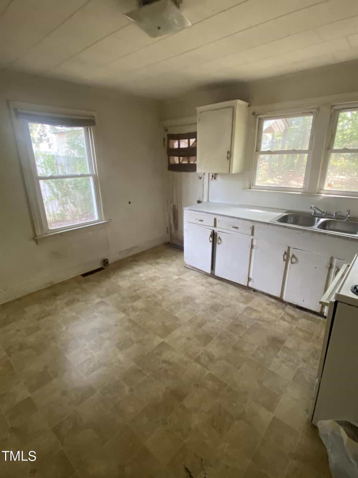 A small, empty kitchen with beige vinyl flooring, white walls, and a ceiling-mounted light fixture. It features white cabinets, a stainless steel sink under double windows, and an old wooden door with a small window. The kitchen appears worn and dated.
