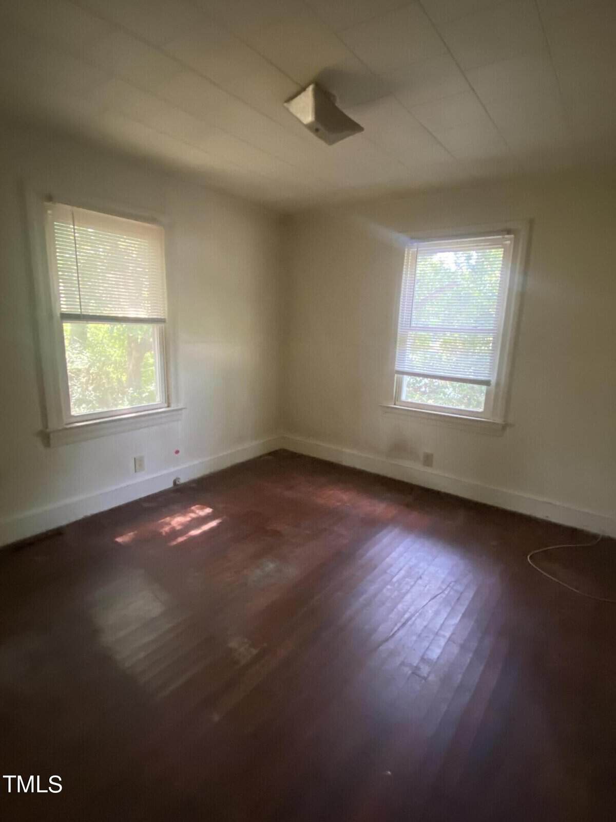 A small, empty room with two windows allowing natural light to enter. The room has plain white walls and a polished hardwood floor. A simple light fixture is mounted on the ceiling. Blinds partially cover the windows, and a few electrical outlets are visible.