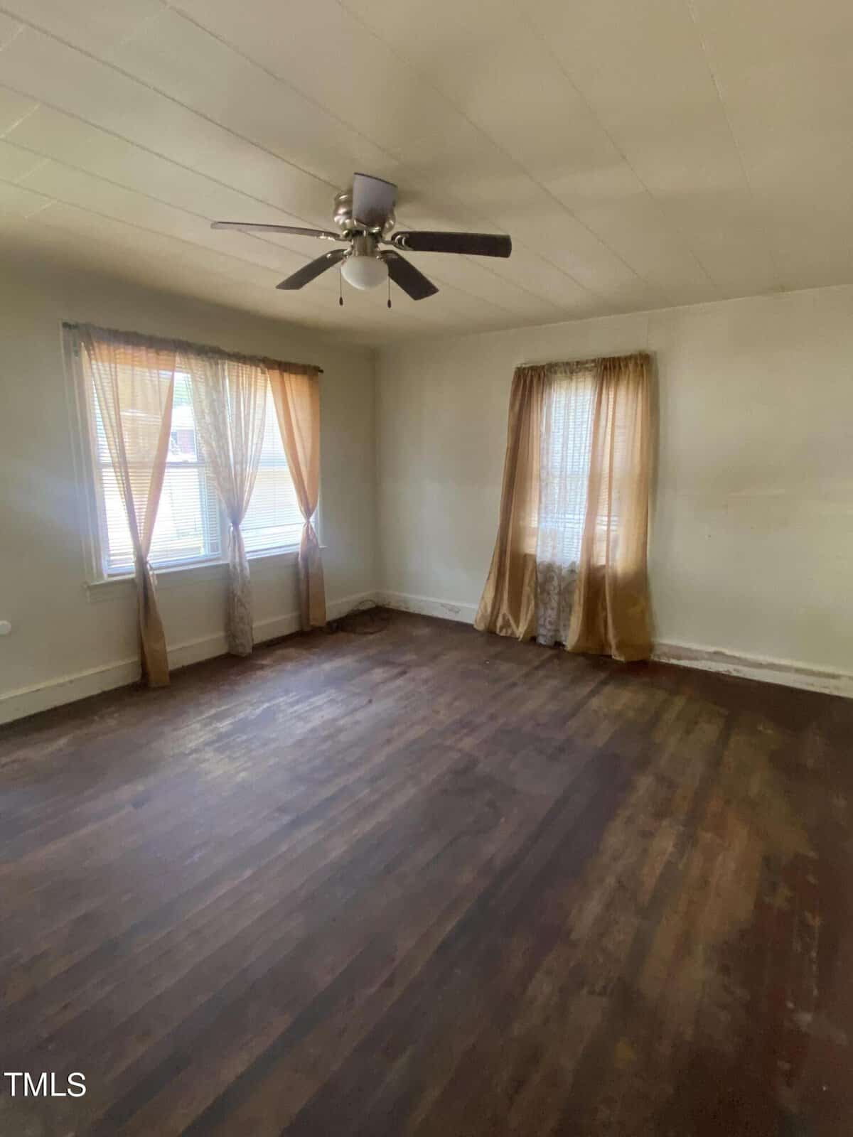 A vacant room with wooden flooring, two windows covered with light brown curtains allowing natural light, and a ceiling fan with light fixture in the center. Walls are plain off-white, and the room appears to be well-lit and spacious.