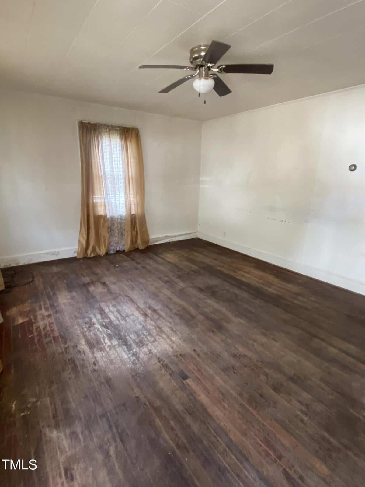 A sparsely furnished room with a single window covered by sheer and opaque curtains. The wooden floor appears worn, and the white walls have subtle marks and discoloration. A ceiling fan with a light fixture is mounted in the center of the ceiling.