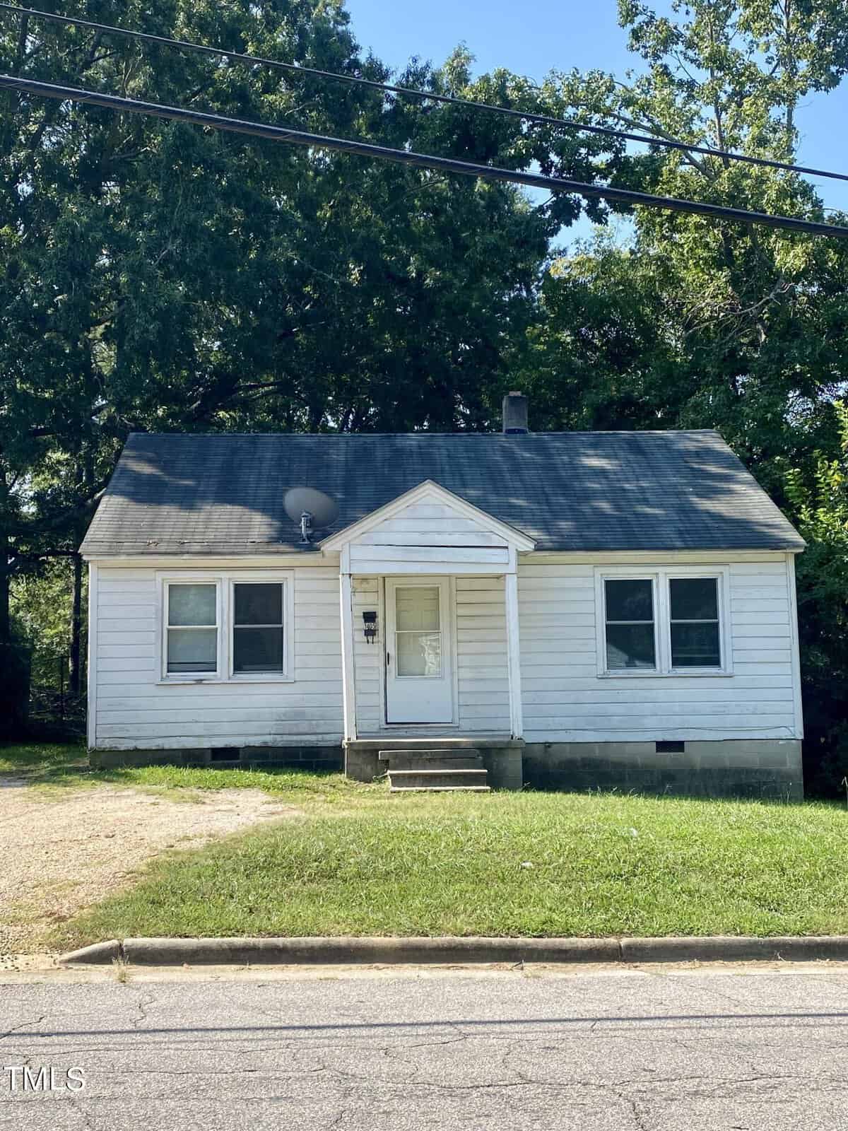 A small, single-story house with white siding and a weathered, dark-gray shingle roof at 1405 Parham Street. There are two front windows and a small covered entryway. Set back from the street in Henderson, it features a narrow lawn and gravel driveway, surrounded by trees.