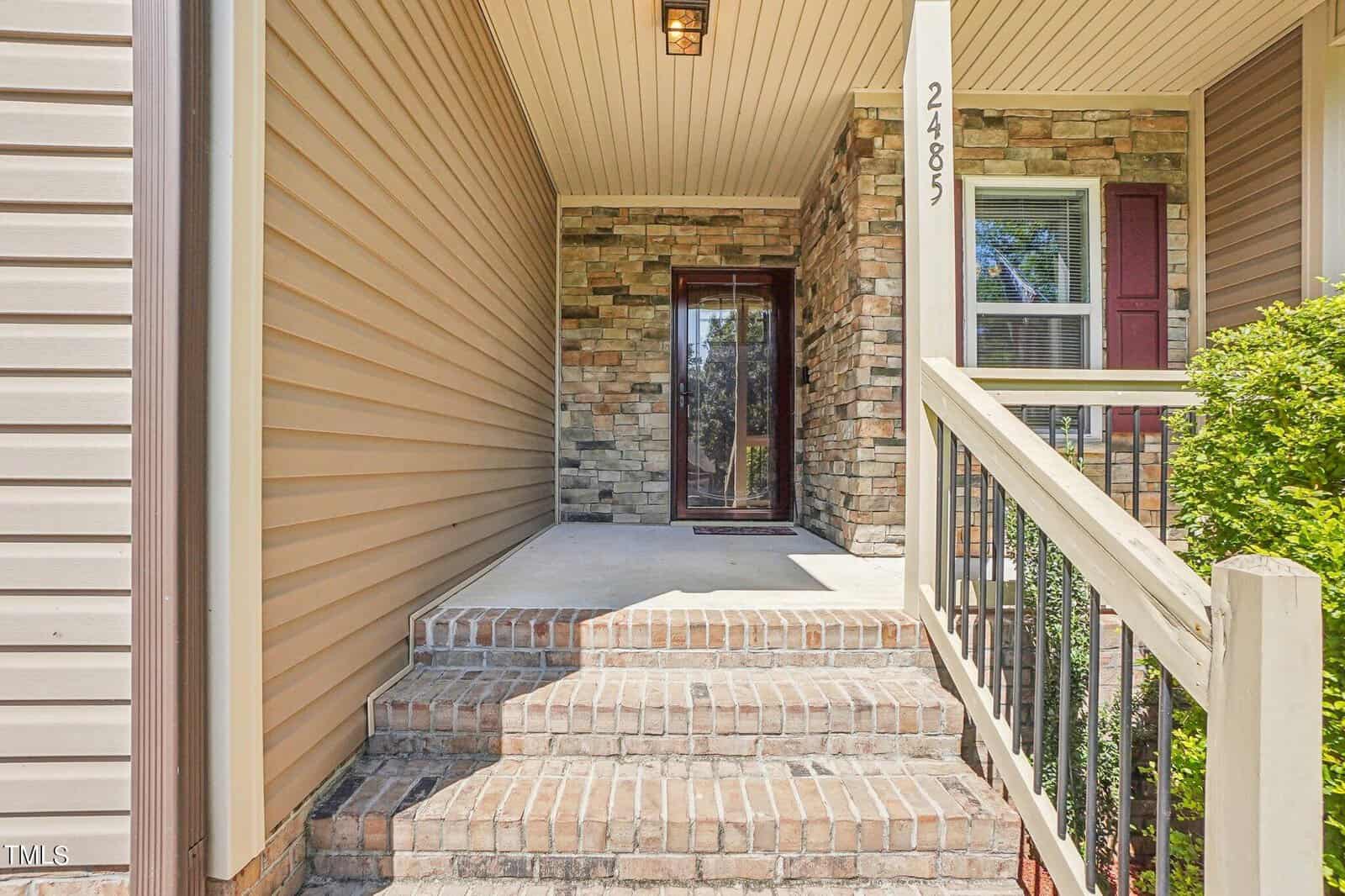 The image shows a house entrance with a covered porch area. The porch is accessed by a few brick steps and has a stone facade. The house number "2485" is displayed on a white column. Bushes are visible beside a window with red shutters.