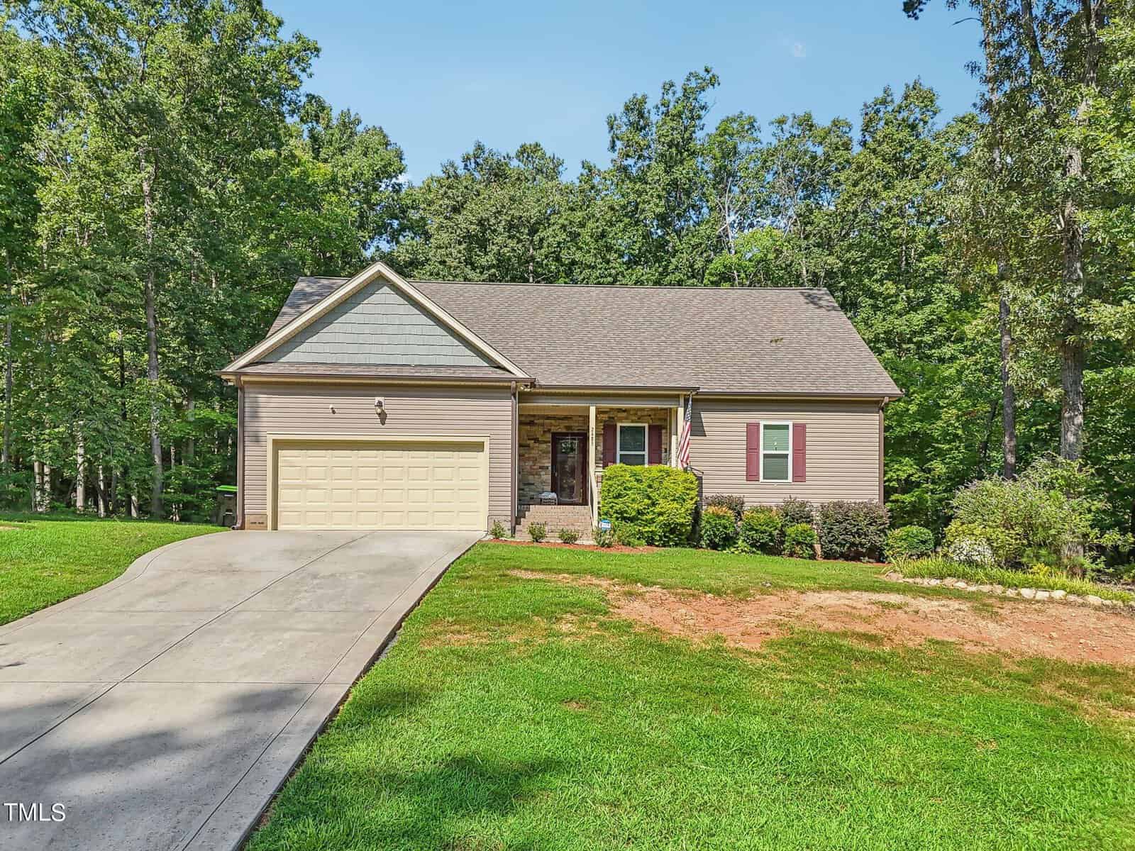 A single-story house with a sloped roof, two-car garage, and front porch. It's surrounded by a neat lawn and trees in the background. An American flag hangs near the porch.