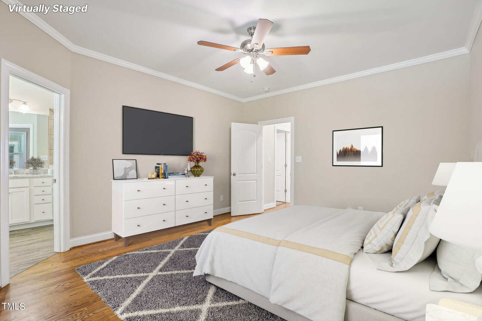 A virtually staged bedroom with a bed, dresser, and TV. The bed has patterned pillows and a beige throw blanket. A ceiling fan is above, and the room features hardwood floors. An open door leads to a bathroom with a visible vanity.