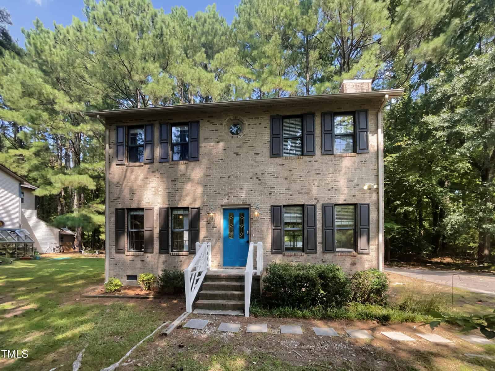 The charming two-story brick house at 612 13th Street in Butner features blue shutters and a matching front door, nestled among trees. A small front porch with steps guides you to the entrance, while a stone path winds through the yard beneath a clear, sunny sky.
