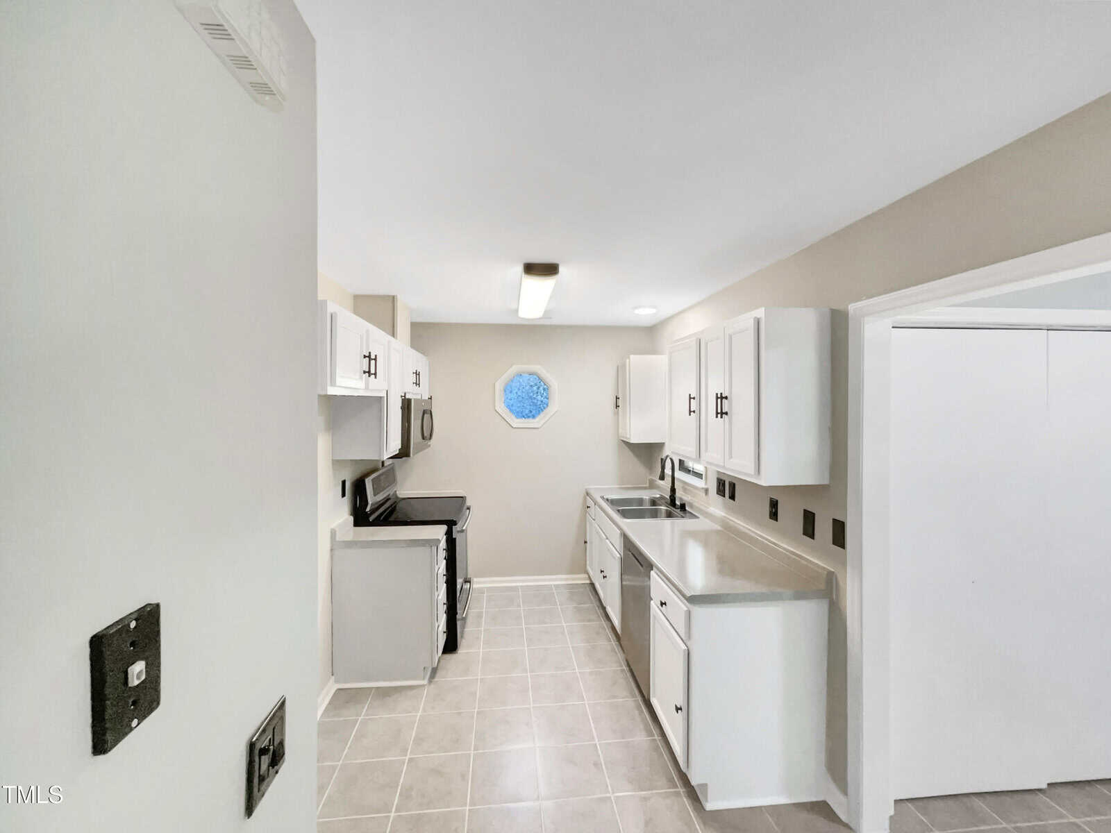 A modern kitchen with white cabinets, stainless steel appliances, and a light gray tile floor. The room features a unique octagonal window above the stove area. The walls and ceiling are painted white, providing a clean and bright atmosphere.
