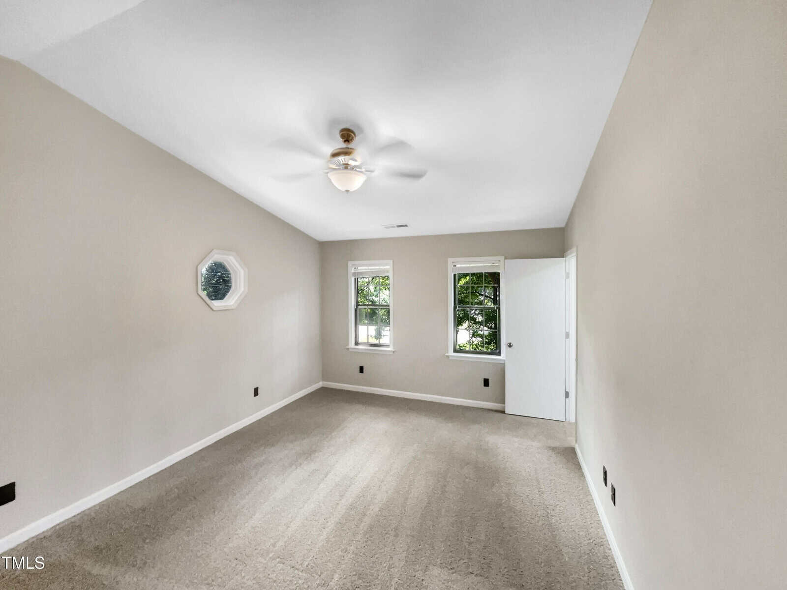 A vacant room with light gray walls and beige carpet. The ceiling features a white fan with a light. There are two rectangular windows with white trim and a small octagonal window on the left wall. An open white door is visible on the right.