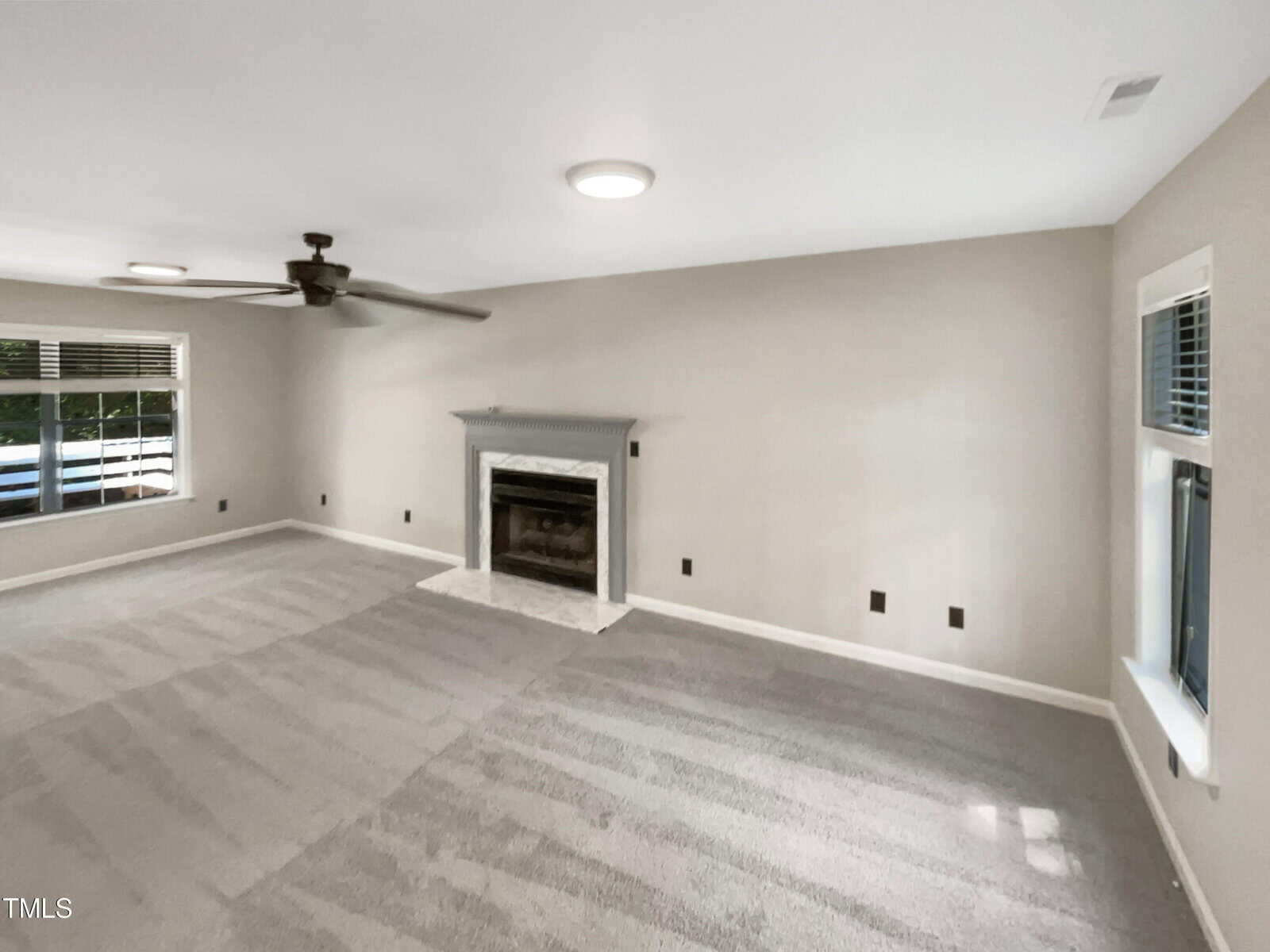 A spacious, unfurnished room with light gray walls and carpeted floor, featuring a fireplace with a white mantel. There is a ceiling fan with light. Two windows with blinds let in natural light.
