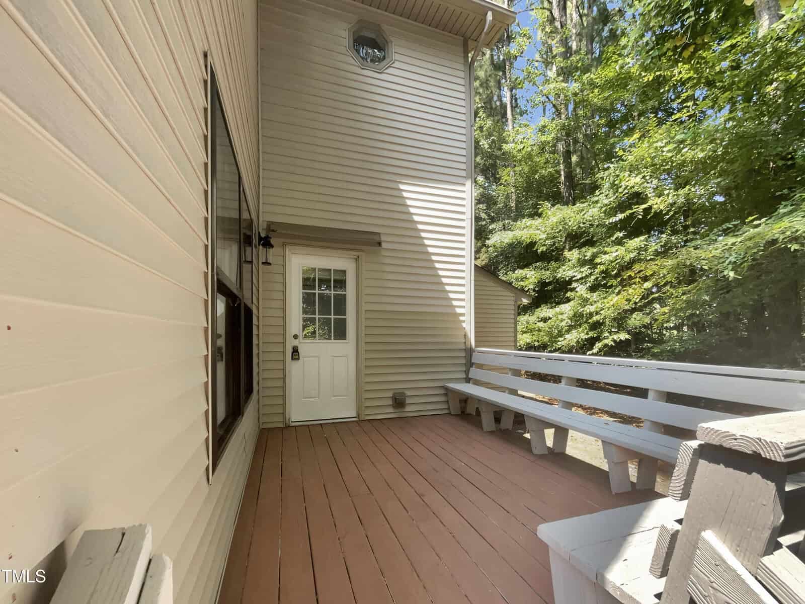 A view of a wooden deck attached to a house with beige siding. The deck has a white bench along the right side and a door leading inside. Trees with green foliage are visible in the background under a clear blue sky.