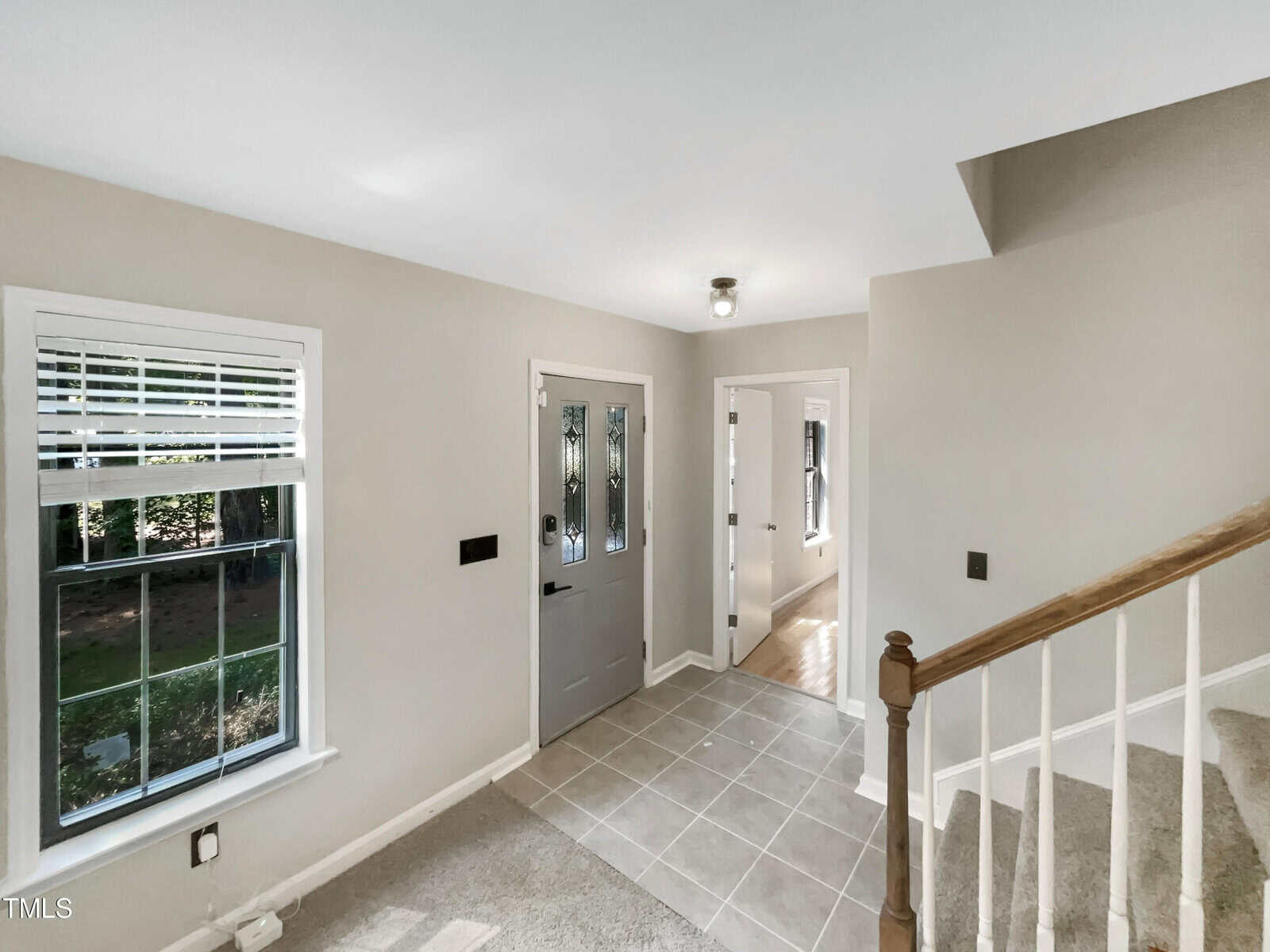 View of a bright, neutral-toned entryway featuring a gray door with a window, white-framed windows on the left, tiled floor, carpeted stairs, and a hallway leading to additional rooms. A ceiling light fixture is visible above.
