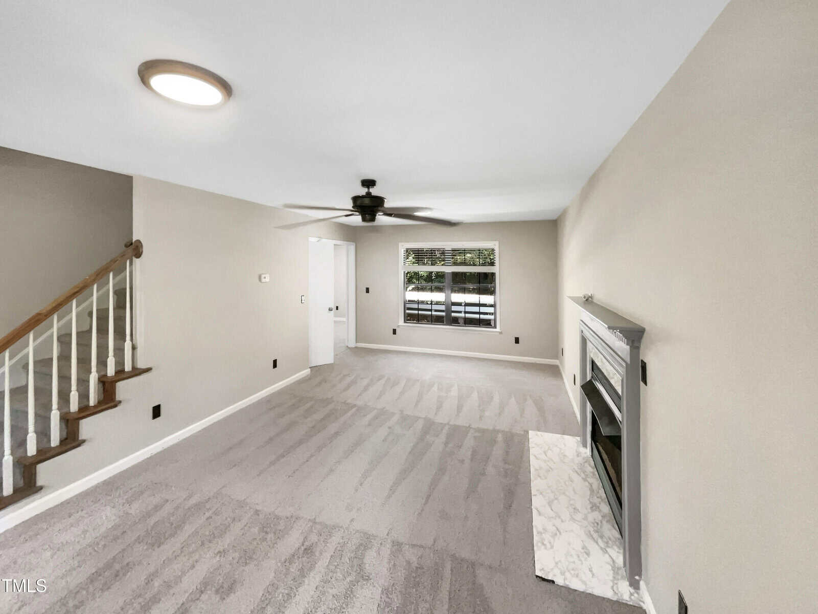 A living room with gray carpet, beige walls, a ceiling fan, and a fireplace with a white mantle. A staircase is on the left side, and a window next to a door is in the background, letting in natural light.