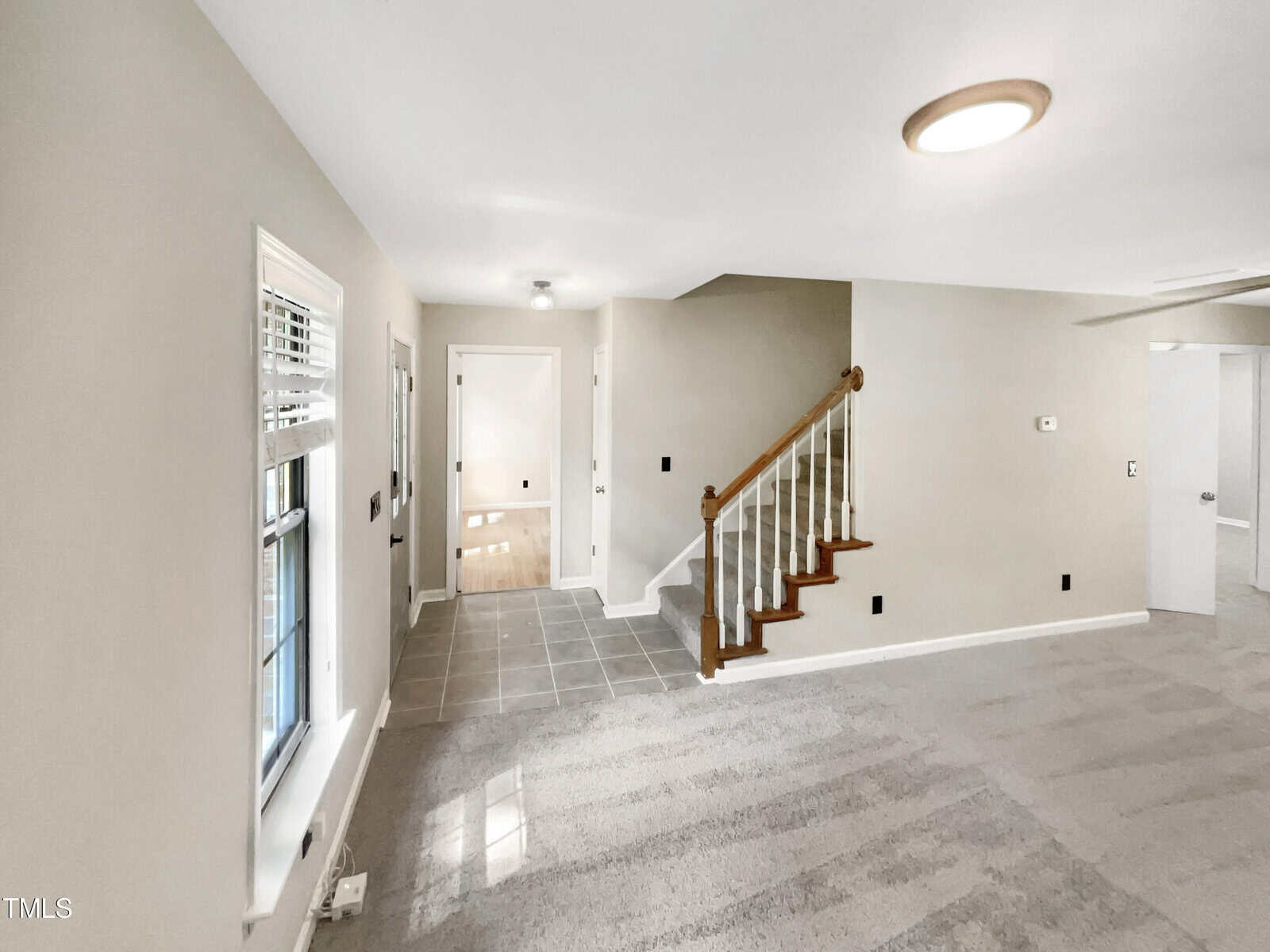 A room with carpeted flooring, stairs with wooden railing leading upward, and a hallway with tile flooring. Sunlight illuminates through a window on the left. The walls are painted light gray, and there is a white door partially open at the end of the hallway.