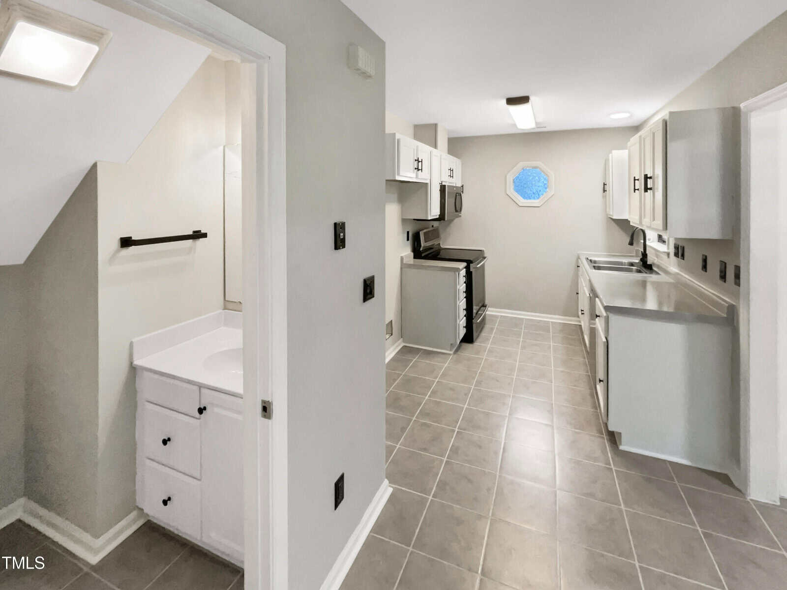 A kitchen with gray tile flooring and white cabinets. It features a stainless steel sink, dishwasher, and stove. There is a small adjacent bathroom with a white sink and cabinet, seen partially to the left. An octagonal window is on the far wall.