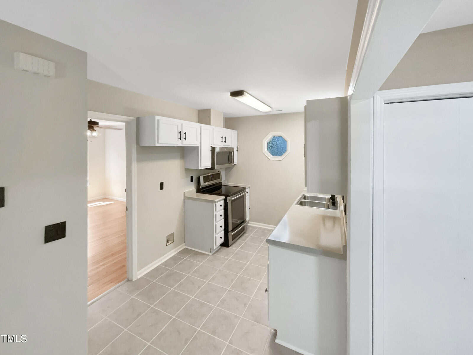 A small kitchen with beige walls features white cabinets, a stainless steel oven, and a dual-basin sink. The floor is tiled in a light color. A unique octagonal window is above the sink. An open doorway leads to another room with wooden flooring.