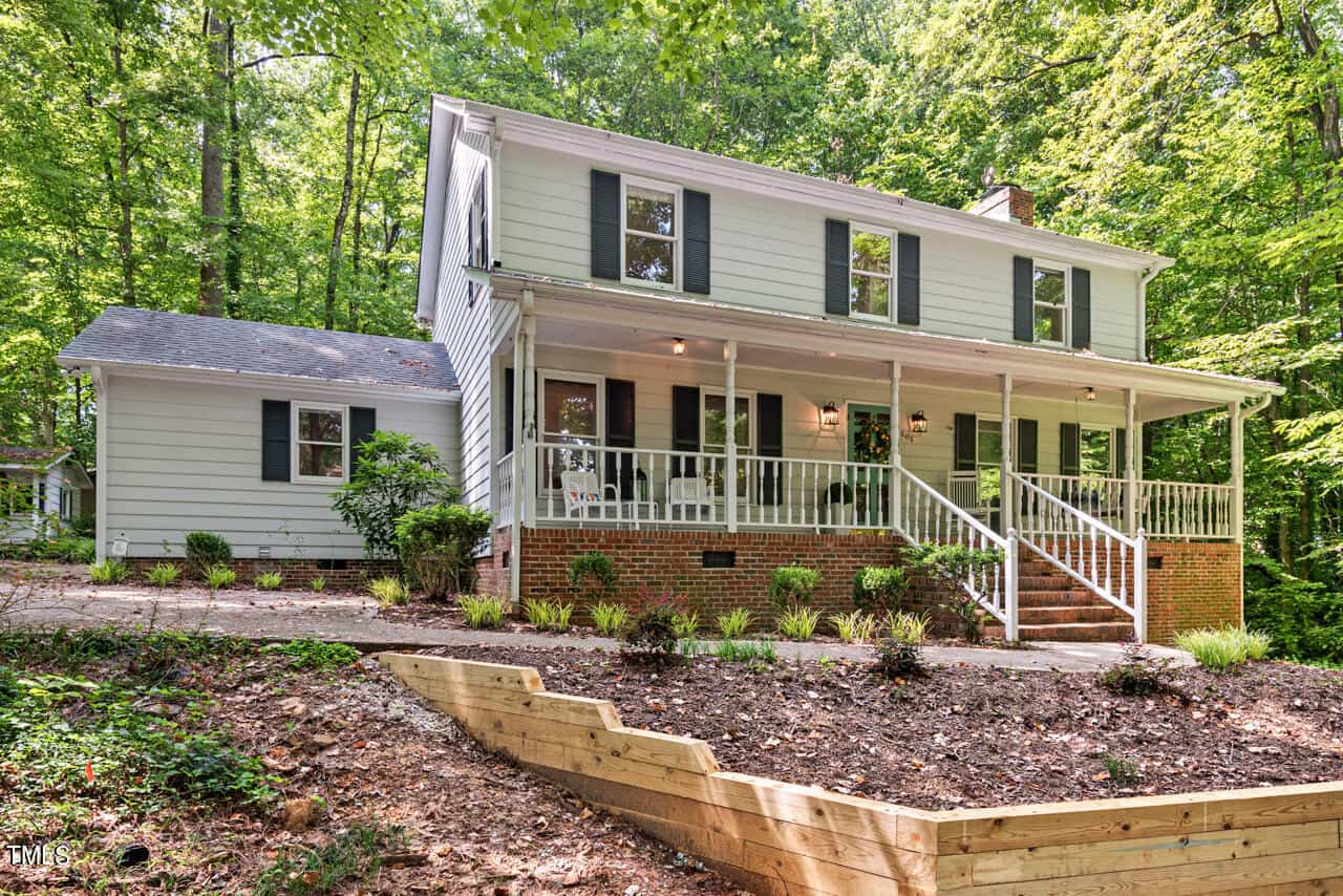 A two-story house with light siding, dark shutters, and a brick foundation. The front has a large porch with white railings and steps leading down to a landscaped yard with a retaining wall. The home is surrounded by lush green trees and shrubs.