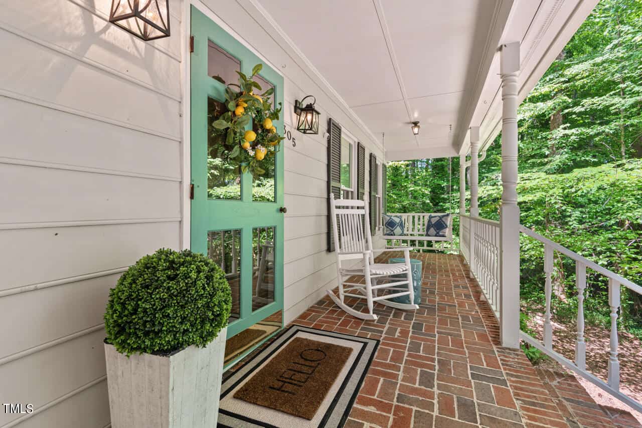 A cozy front porch with a brick floor, featuring a green front door adorned with a lemon wreath. There are two white rocking chairs next to the door, with potted plants and a doormat that reads "HELLO." The porch is lined with white railing and surrounded by greenery.